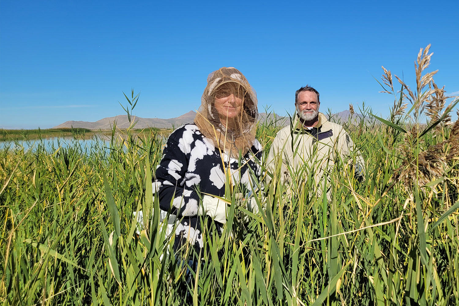 Two adults stand in thick, wait-high vegetation. They are smiling at the camera. One is wearing a bug net.