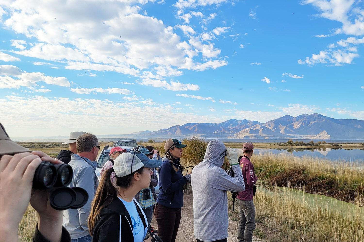 A group of adults look out over a lake with mountains in the distance. Some of them have binoculars.