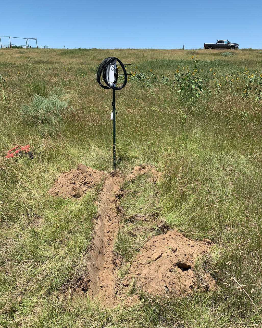 A data logger on a fence post in a prairie with disturbed soil before it.