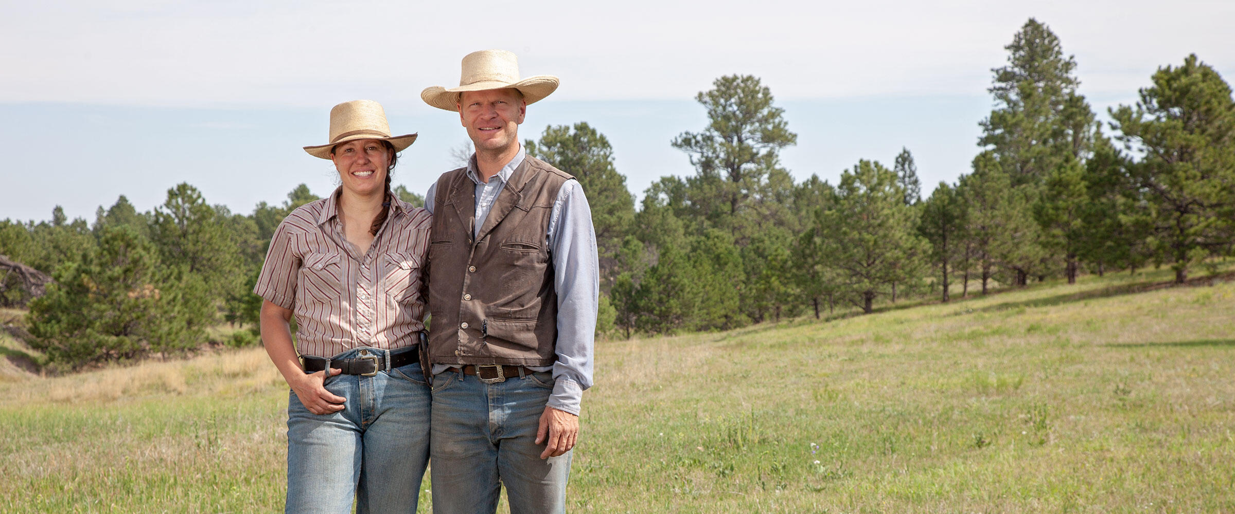 Two ranchers stand in a meadow before in front of pine trees.