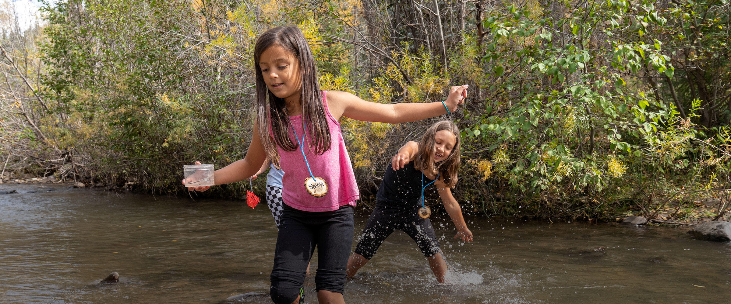 Two girls walk through a stream carrying a jar with an insect.