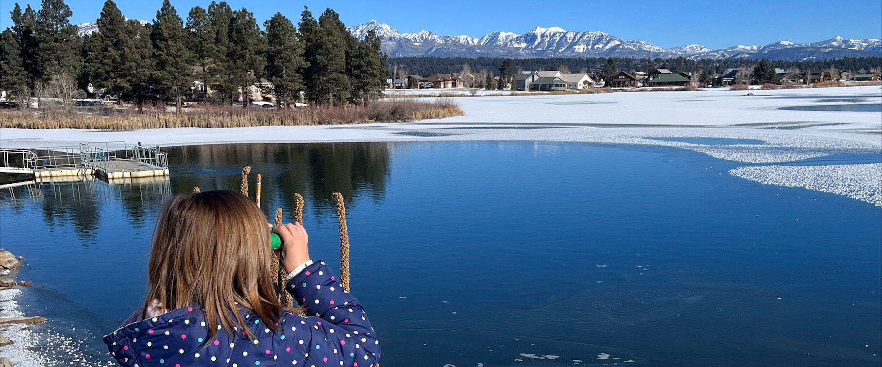 A girl stands in front of an icy lake, looking with binoculars.