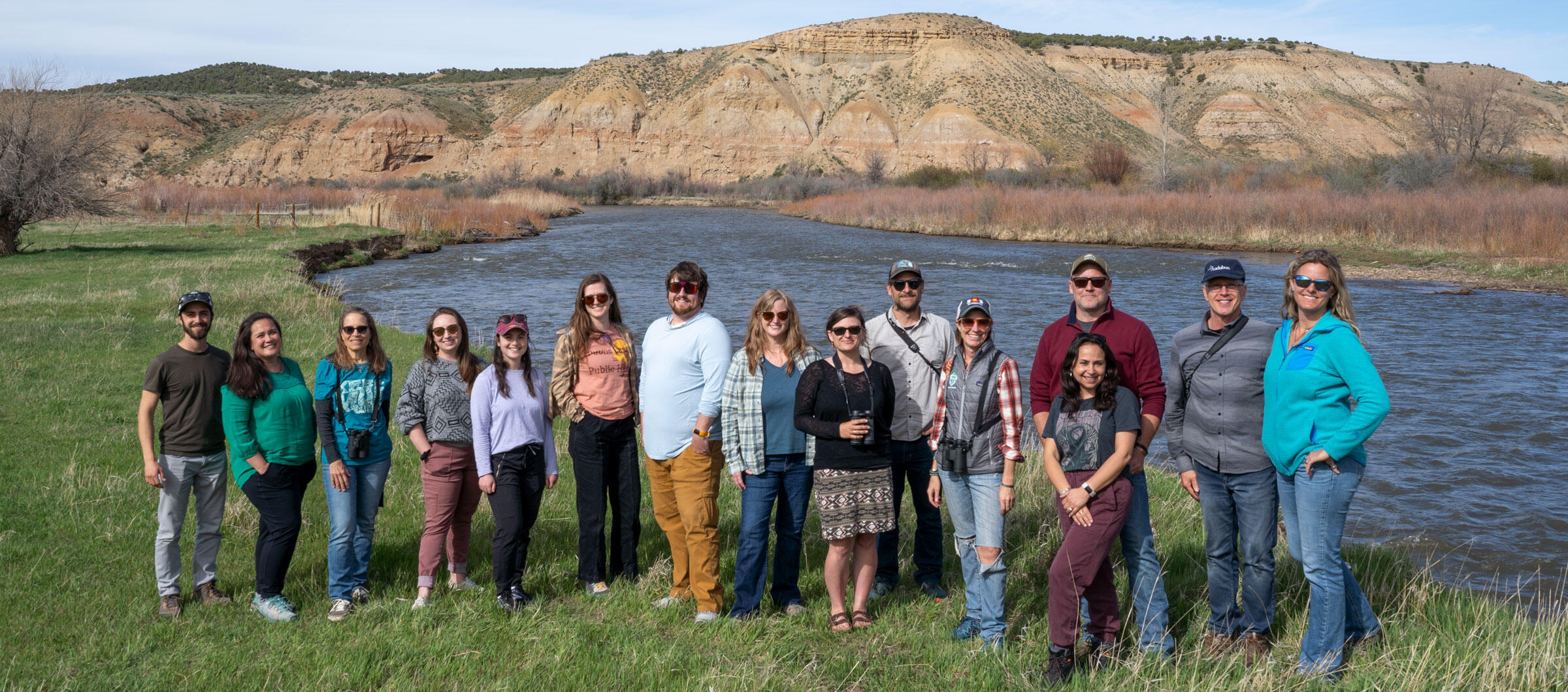 A group of people stand in a field in front of a river and bluff.