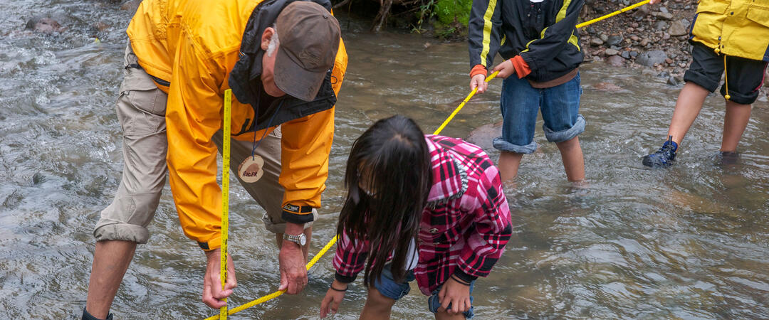 A man helps a group of children take measurements in a river.