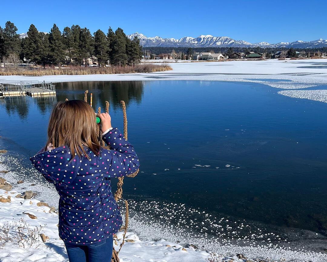 A girl in winter clothes stands in front of a pond, looking with binoculars.