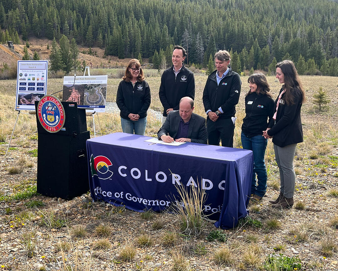 People stand behind Governor Polis as he signs a bill in a montane meadow.